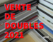 Sur un fond composé d'une photo de style close up, deux piles de livres forme un motif ligné en coin, alternant entre le blanc des pages et les couleurs des couvertures. En premier plan, occupant la partie gauche du montage visuel, nous pouvons lire "Vente de doubles 2021" écrit en rouge vif, doublé d'un violet clair pour créer un effet d'optique