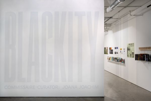 Photo d’une vue de l’exposition Blackity. À gauche de la photo, sur un mur blanc, nous pouvons y lire en gris clair le titre de l’exposition : « BLACKITY » et juste en dessous, en plus petits caractères, « commissaire/curator : Joana Joachim ». À droite de ce mur blanc se trouve une ouverture agissant comme l’entrée de l’exposition. Cette photo nous offre donc un avant-goût de ce que contient l’exposition, l’intérieur de la salle étant que partiellement visible à travers l’entrée de l’exposition.