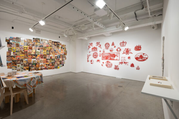 Panoramic view of Manon Tourigny's "Setting the Table" exhibition in Artexte's exhibition space, with its white walls and grey concrete floor. On the left is a partial view of a round wooden table covered with Folie culture's colourful tablecloth. In the centre of the photo, a white wall is occupied by the all-over work "Vieux buffet" by Marc-Antoine K. Phaneuf, consisting of 300 vintage recipe books. To its right, Chef Lee's Wok'n'Roll Garden by Karen Tam marks the white wall with stereotypical Chinese symbols made of finely cut red paper. To the far right, Graff Dinner, with its box and recipes printed on square paper, can be seen on a white shelf on the wall.