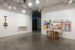 Photo of a partial exhibition view of "Setting the Table", which on the left shows Pierre Ayot's mural "La cuisine de Bocuse" (1986) and the collective work "Graff Dinner" (1978) on a white table to the right of the first. To the right of the image is a view of the Folie Culture themed table. The white walls and grey concrete floor of the exhibition space take up most of the picture.