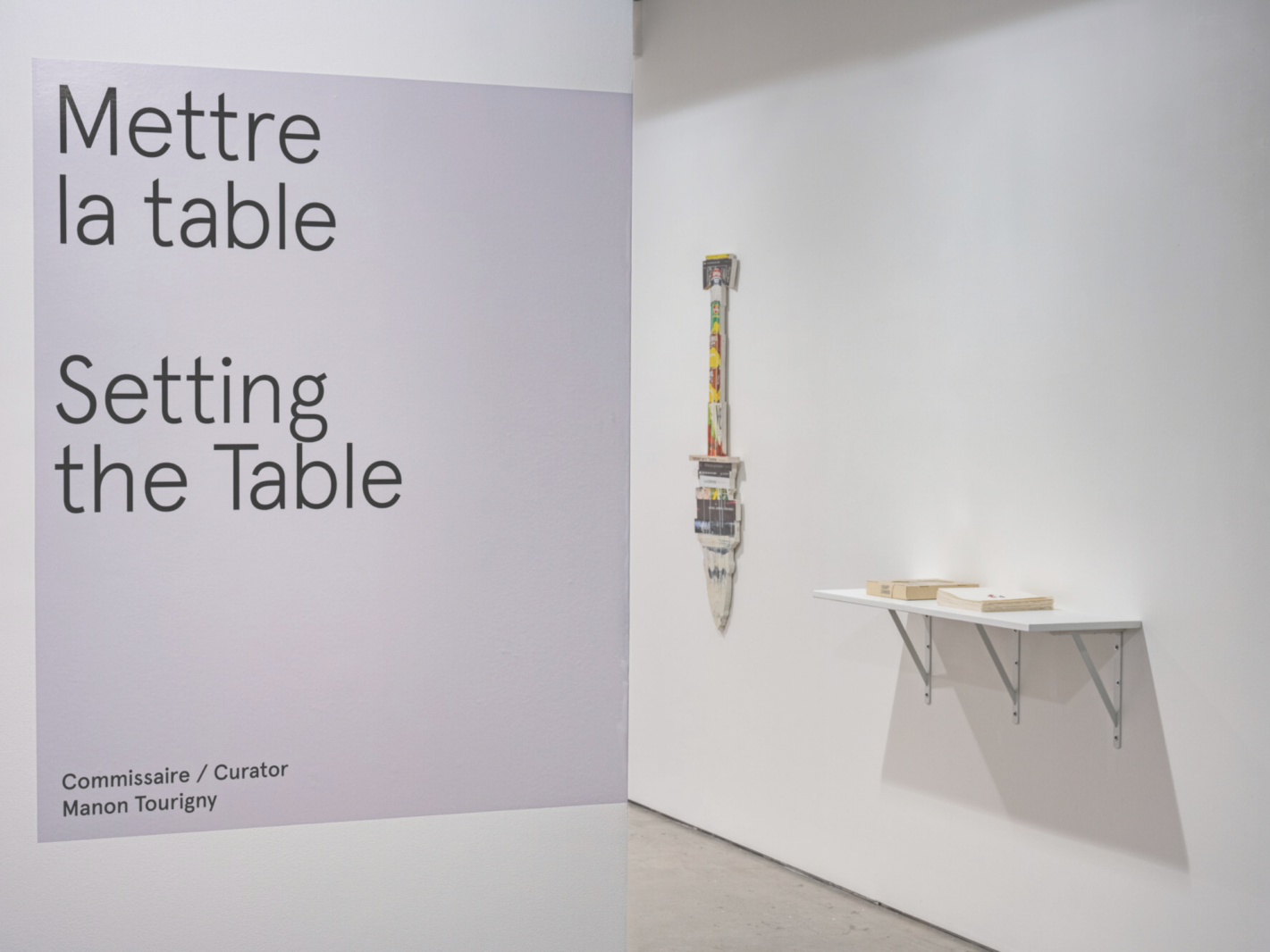 Landscape photography of the entrance to Artexte’s white exhibition space. On the wall to the left of the photo is the title of the exhibition “Mettre la table. Setting the Table. Commissaire/Curator Manon Tourigny” on a grey vinyl. The exhibition opens on the right. Pierre Ayot’s “Graff Dinner” is displayed on a white shelf. Further back, the works “La cuisine de Bocuse” by the same artist is presented on the wall.