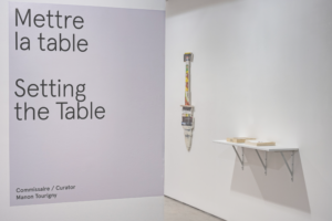 Landscape photography of the entrance to Artexte’s white exhibition space. On the wall to the left of the photo is the title of the exhibition “Mettre la table. Setting the Table. Commissaire/Curator Manon Tourigny” on a grey vinyl. The exhibition opens on the right. Pierre Ayot’s “Graff Dinner” is displayed on a white shelf. Further back, the works “La cuisine de Bocuse” by the same artist is presented on the wall.