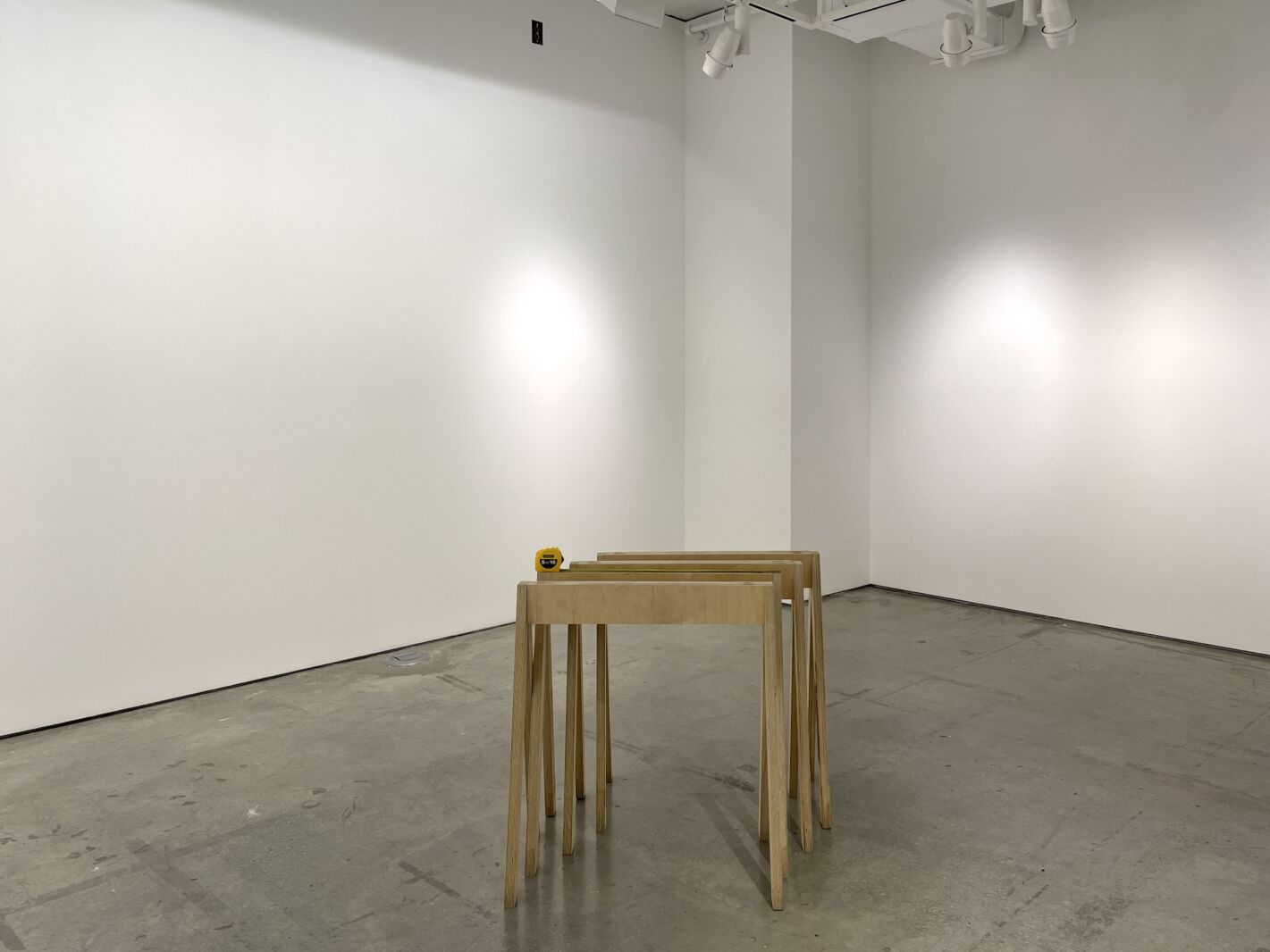 The photo shows a partial view of the exhibition space with white walls and light grey concrete floor. In the center of the photo are a series of four wooden display case supports, set one into the other. A yellow measuring tape rests on the second one.