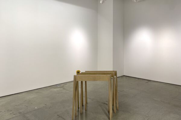 The photo shows a partial view of the exhibition space with white walls and light grey concrete floor. In the center of the photo are a series of four wooden display case supports, set one into the other. A yellow measuring tape rests on the second one.
