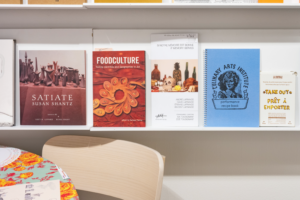 Landscape colour photo showing a detail from the "Setting the Table" exhibition. White shelves occupy the white wall of the Artexte exhibition room. Several coloured documents and publications are visible, or partially visible. On the left, in the foreground of the photo, the back of a light-coloured wooden chair is visible, alongside a partially visible table covered in a tablecloth coloured blue with orange and red motifs.