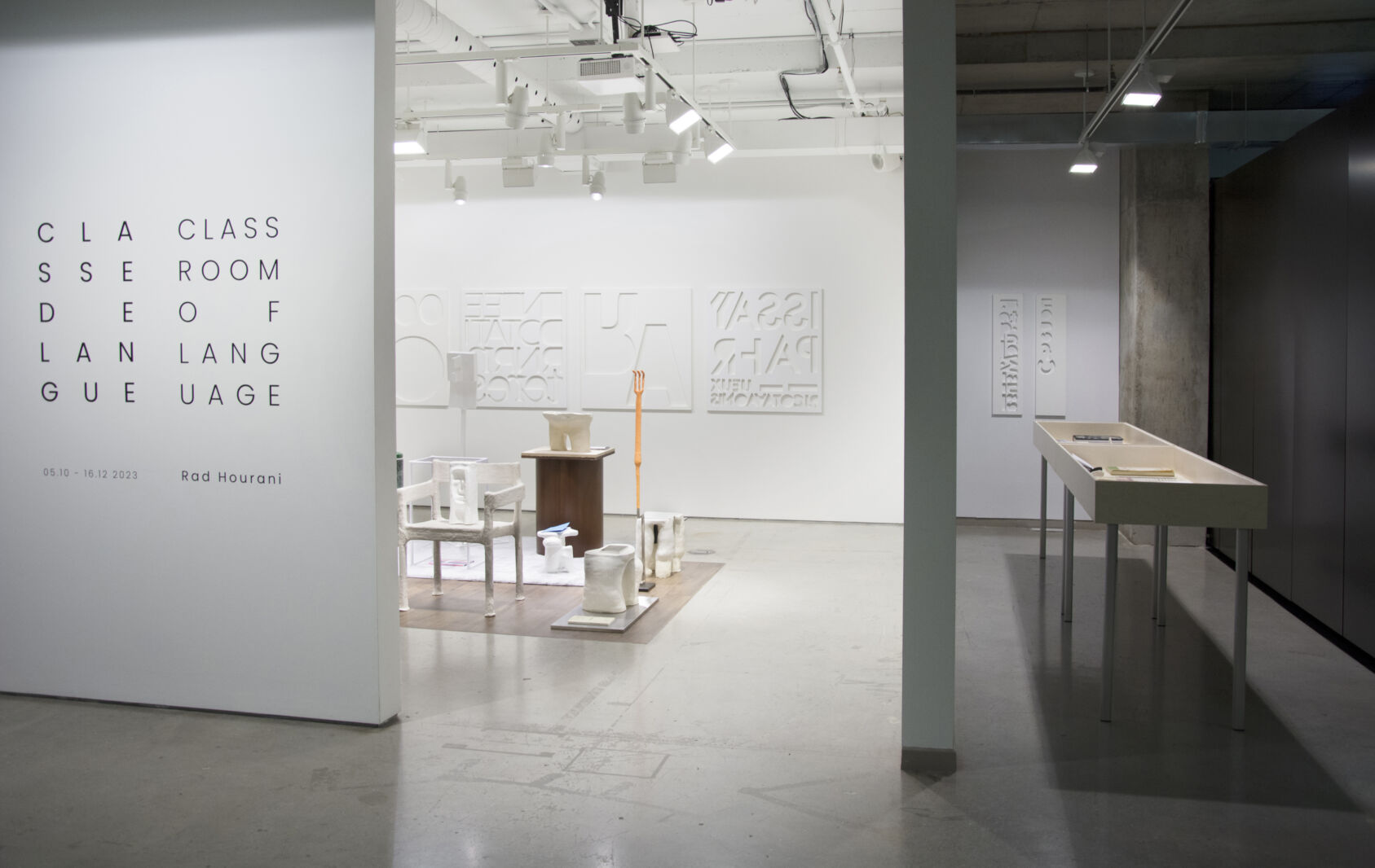 Photographie couleur de format paysage d'une vue générale de l'exposition « Classe de langue » de Rad Hourani. Le titre de l'exposition est sur le mur extérieur, et on peut apercevoir au centre de la salle des sculptures de différents formats et matériaux. Sur les murs se trouvent des oeuvres blanches où sont découpées des formes.