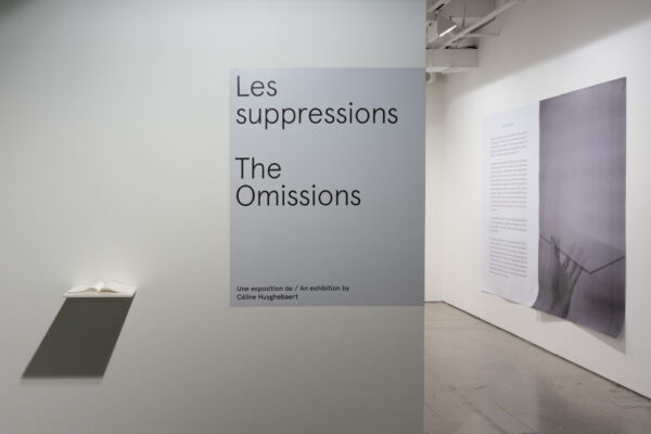 Photographie couleur format paysage de l'entrée de l'exposition « Les suppressions ». On y aperçoit le titre de l'exposition imprimé en grands caractères sur le mur d'entrée, avec une petite tablette avec un livre blanc. On peut apercevoir un mur à l'intérieur de la salle, sur lequel est affichée une page de livre imprimée en très grand format.