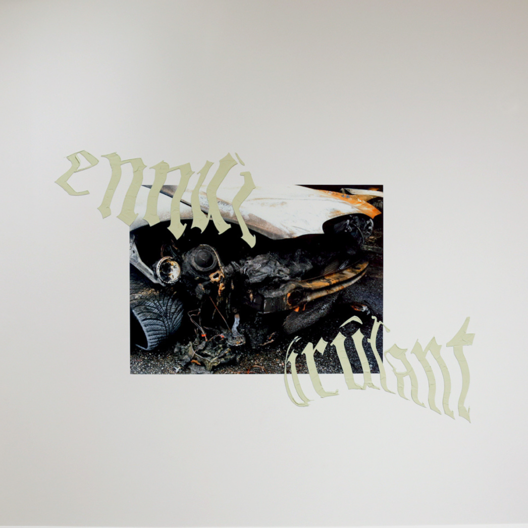 Photographie couleur format carré d'une oeuvre installée sur un mur blanc. On peut y lire « ennui brûlant » en typographie gothique autour d'une photographie du pare-choc d'une auto ayant l'air brûlé.