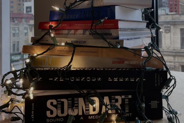 Square colour photograph showing a pile of books of varied formats and colours on a table. A garland of lights is placed on top of the books in a decorative manner.