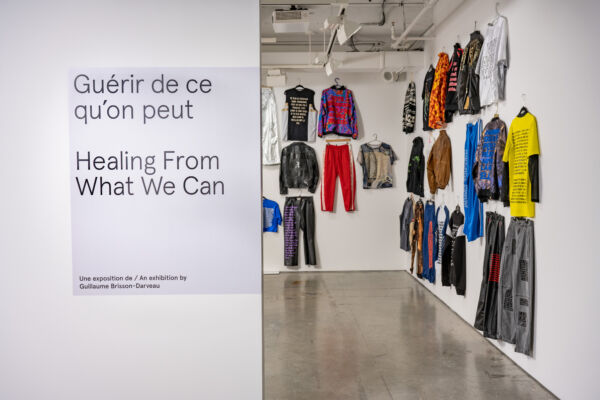 Colour photograph showing the entrance to the "Healing from what we can" exhibition. The title of the exhibition in French and English is on the left outside wall, and we can see inside the exhibition room a number of colourful clothes hung across the walls.
