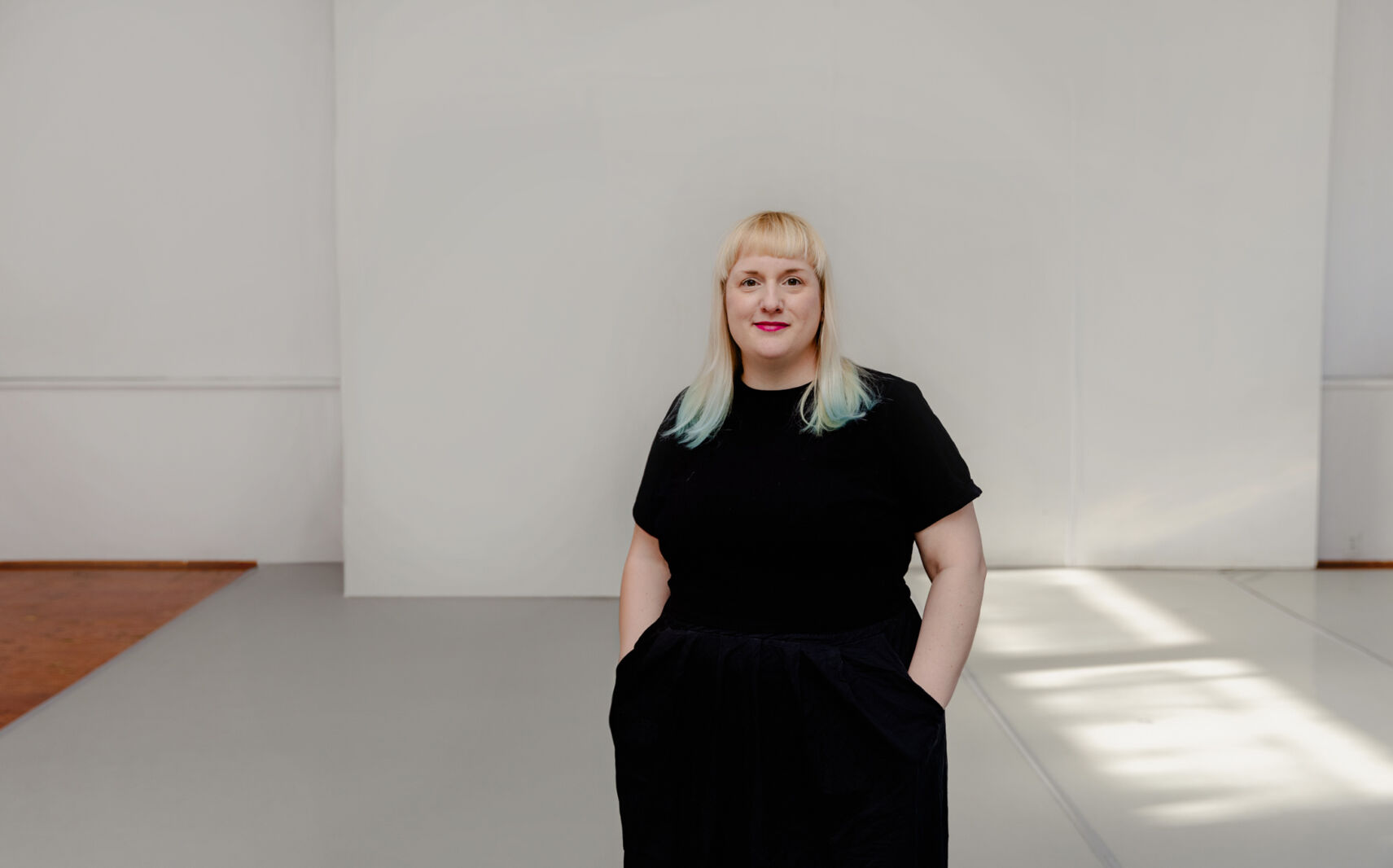 Colour photograph showing a person wearing a black t-shirt and black pants standing in front of a white wall. She has long blond hair with blue tips, and is standing with her hands in her pockets.