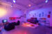 Colour photograph showing the inside of an exhibition where the light is dimmed and tainted purple and orange. Two beanbag chairs are set up in the left corner in front of a TV, while a larger grey couch is placed upon the right wall, in front of a beige carpet and many pillows. Posters are placed alongside the right-side wall.