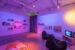 Colour photograph showing the inside of an exhibition where the lights are dimmed and tainted purple and orange. We see two beanbag chairs on the right side, with shelves showing magazines behind them. On the left wall are many shelves showing artist documents and books. The entrance to the exhibition is in the centre, covered by a grey curtain.