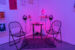 Colour photograph showing a close-up of a corner in an exhibition, where the lights are dimmed and tainted purple and orange. We see two black metallic chairs and a small table, on which documents are placed. Besides each chair is a little shelf on which an MP3 player and headphones are attached. There is also a lava lamp on the right side.