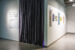 Colour photograph showing the entrance to an exhibition. The title of the exhibition is printed on a vinyl on the left side, while posters are placed up on the right wall. The entrance is in the middle, covered by a large grey curtain.