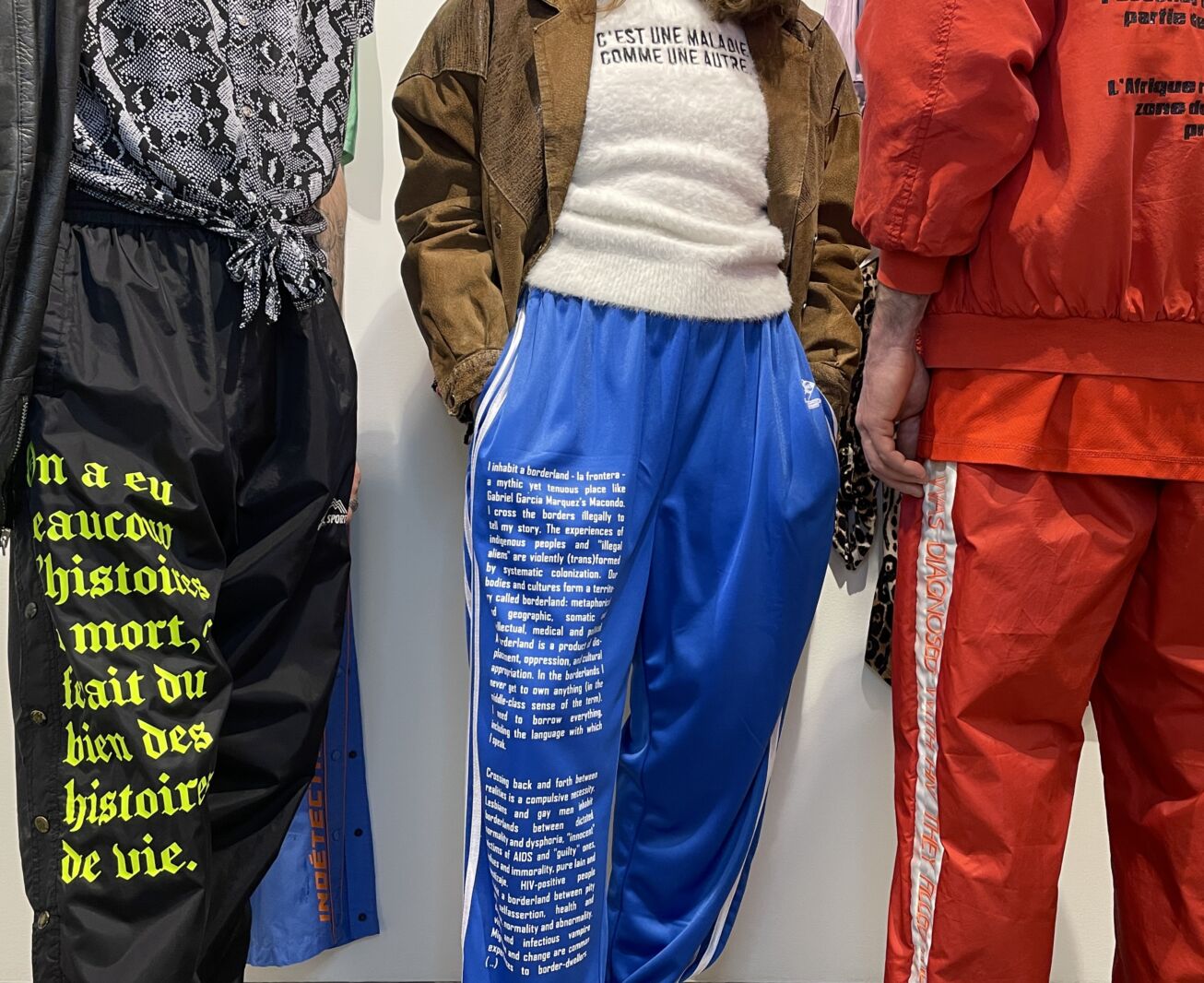 Colour photograph showing the bodies of three people wearing clothing inspired by the 1980s and 1990s, on which quotations have been printed in connection with the AIDS/HIV crisis.