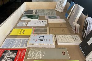A square-format colour photograph on which a rectangular display case made of light-coloured wood holds a variety of carefully placed documents in a range of colours and formats.