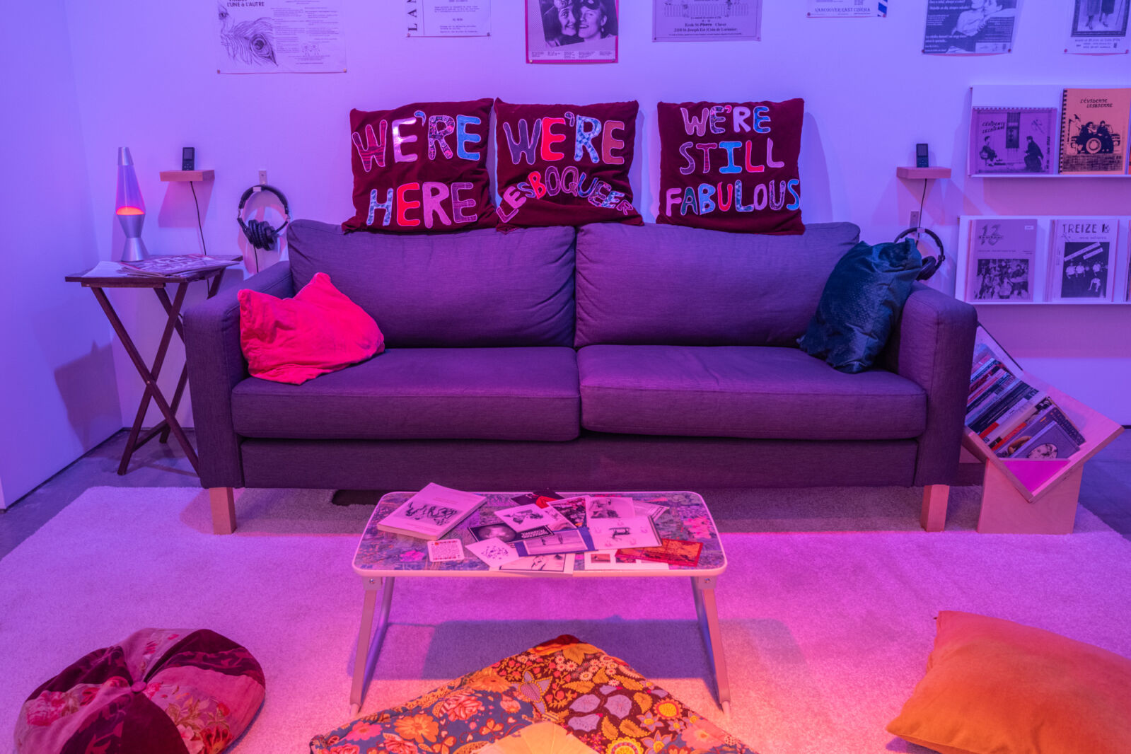 Colour photograph of an exhibition room made to look like a living room. There is a grey couch with pillows placed on top that say "We're Here" "We're Lesboqueer" and "We're Still Fabulous". There is a coffee table in front of the couch, placed on top of a beige rug, where other pillows are laid out. There are posters on the wall. The lighting in the room is dimmed and tainted purple and orange.