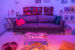Colour photograph of an exhibition room made to look like a living room. There is a grey couch with pillows placed on top that say "We're Here" "We're Lesboqueer" and "We're Still Fabulous". There is a coffee table in front of the couch, placed on top of a beige rug, where other pillows are laid out. There are posters on the wall. The lighting in the room is dimmed and tainted purple and orange.