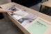 Square color photograph of a wooden display case. It shows three documents bound in red spiral - one with green paper, on which the title of the project is printed, and two others to its left. The farthest displays a black-and-white photograph, and the other, text. A hand can be seen turning the page of the document in the center of the display case. An image of a black-and-white duvet is affixed to the bottom left-hand side of the display case.