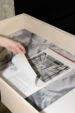 Colour photograph showing a spiral-bound document placed on a large scale image of a duvet cover inside a wooden display case. A hand is flipping through the document.