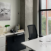 Colour photography showing an office. We see a desk and black chair in the forefront of the image, with a small bookshelf and cabinet in the back. On top of the shelf and cabinet are files, a lamp and a phone. There is a black and white photograph hanging on the wall.